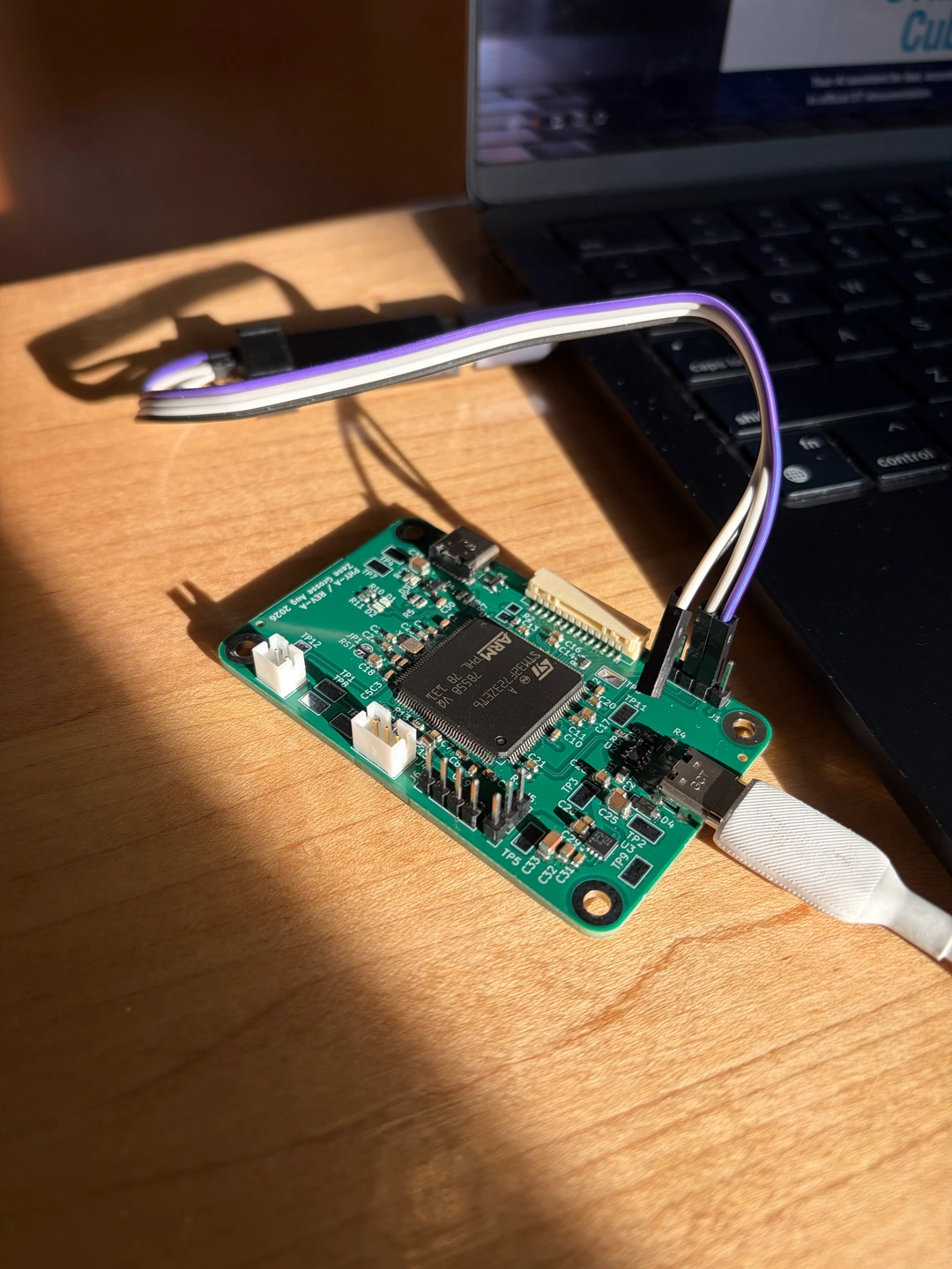 An STM32F723 board being programmed beside a laptop, with programming leads and a USB-C cable connected, illuminated by warm sunlight.